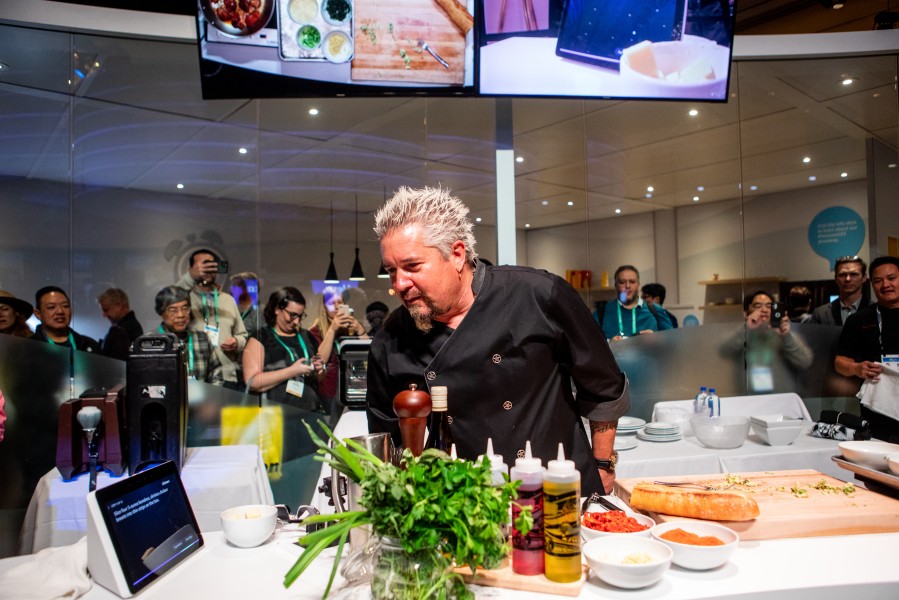 Guy Fieri prepares a sandwich for onlookers behind a glass wall.