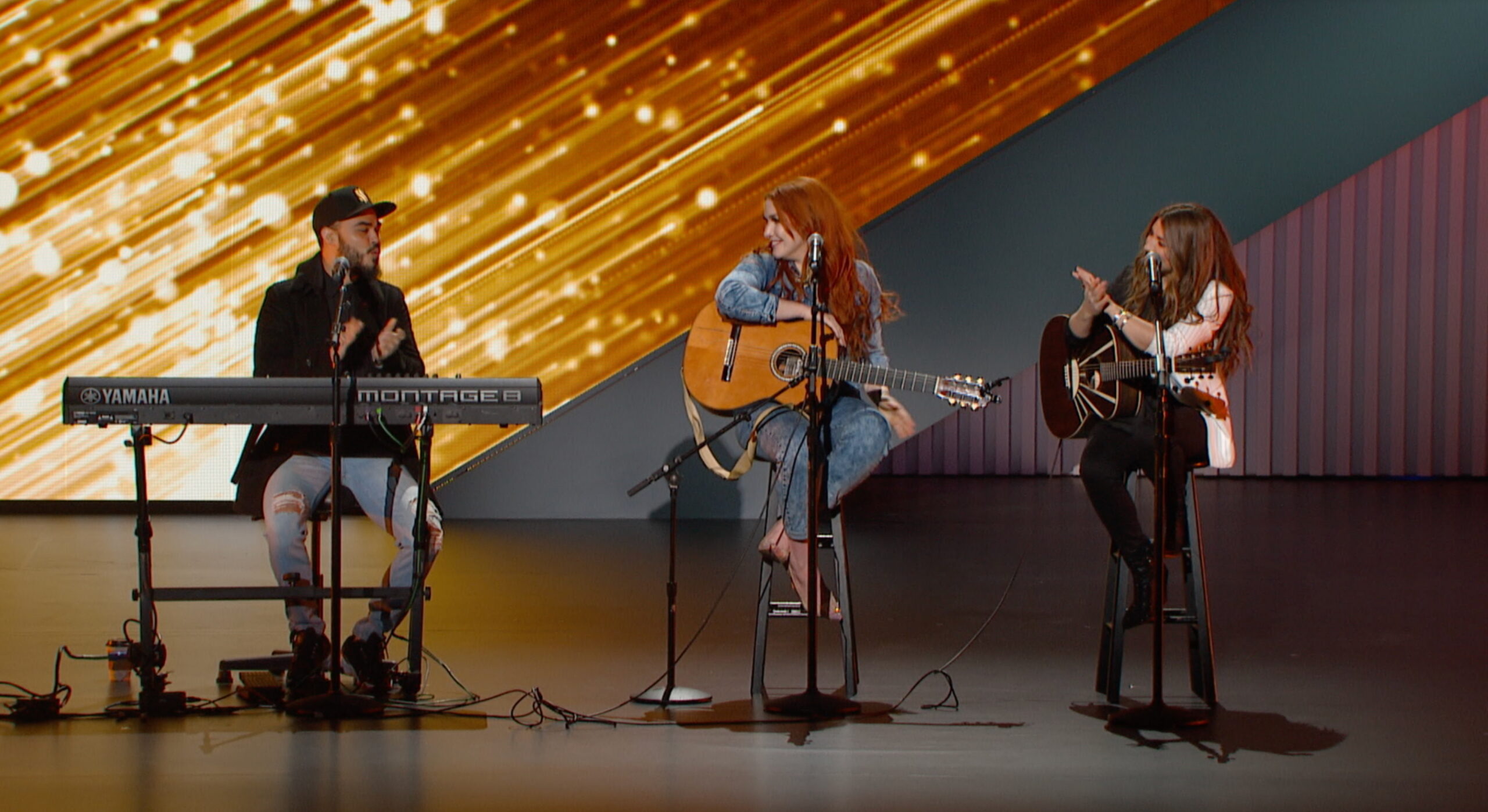 Group of three musicians on a stage in a songwriters' round fashion with a keyboard and two guitars